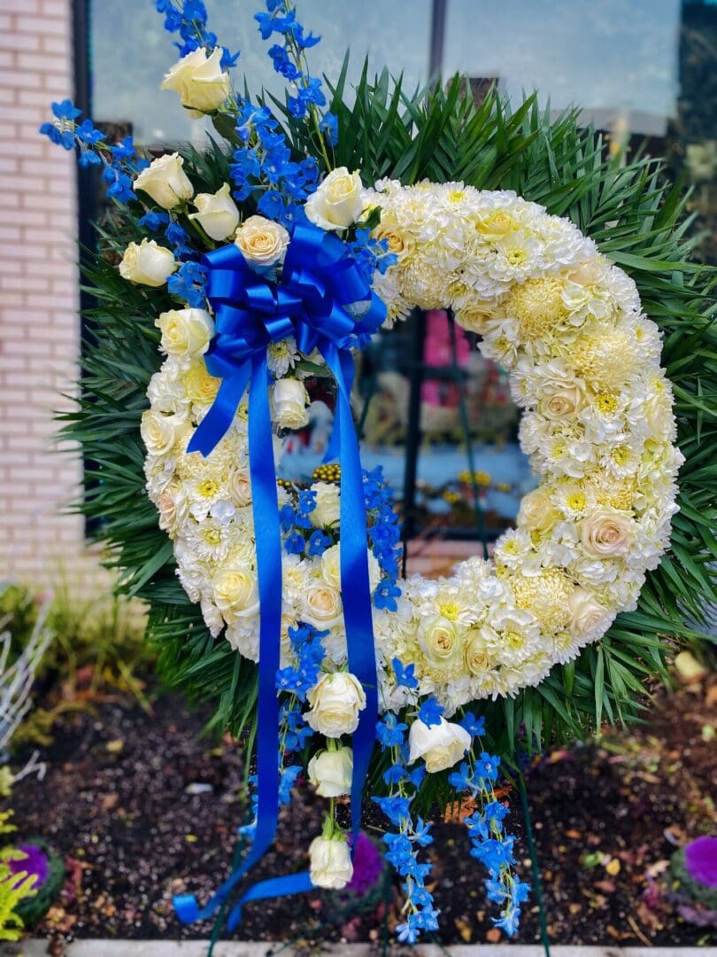 A BLUE MOON WREATH SPRAY with white and yellow flowers, featuring blue accents and ribbons, displayed on a stand.
