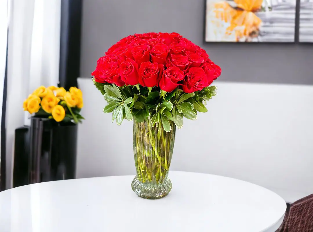 A vibrant bouquet of 50 Premium Red Long Stems Roses in a clear vase on a white table with a blurred background.