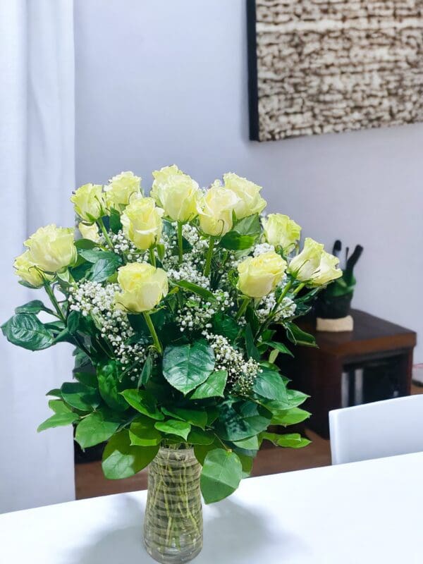 A vase of 24 white premium long stems roses near baby's breath on a table in a room with white walls and decorative elements.