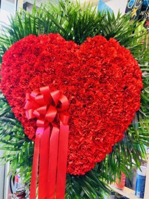 Large bleeding heart red carnations floral arrangement with a pink ribbon, set against green leaves.