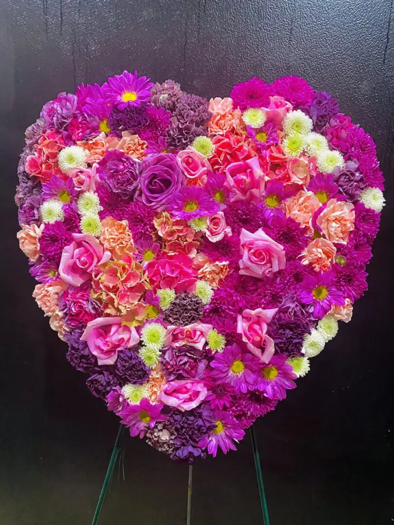 A vibrant Lavender Love Heart Standing Spray with various shades of pink and purple flowers displayed against a dark background.