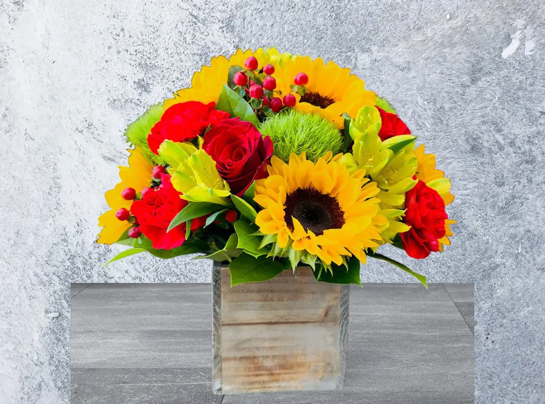 A vibrant bouquet with sunflowers, 12 PAYASITOS ROSES, and greenery in a rustic wooden vase against a textured grey background.