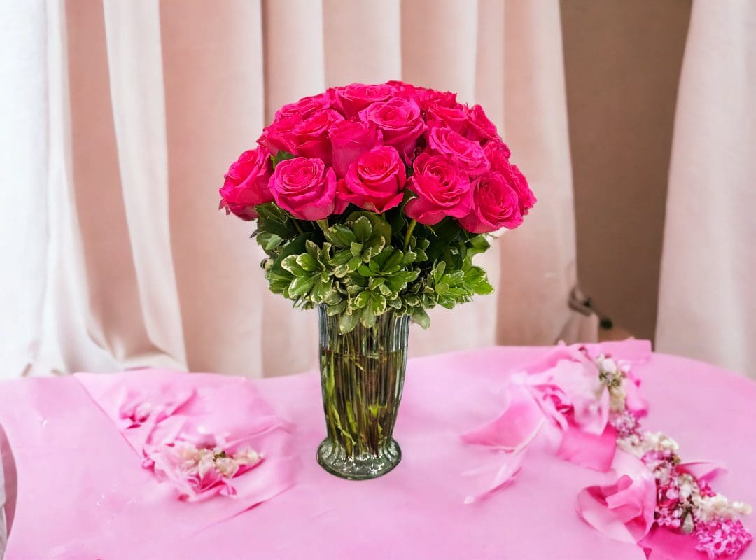 Bouquet of 12 PAYASITOS ROSES IN VASE arranged in a clear vase on a table with a pink cloth and scattered petals.