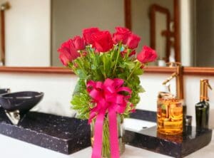 A bouquet of 12 PAYASITOS ROSES IN VASE with a pink ribbon on a bathroom counter.