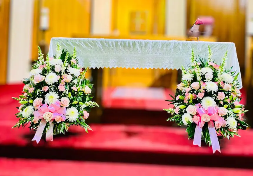 Two floral arrangements flank a white-draped altar in a church setting.