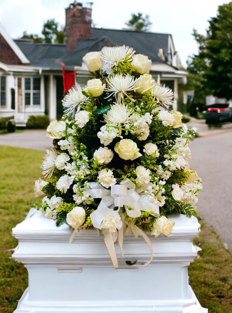 Floral arrangement atop a white casket outside a house.