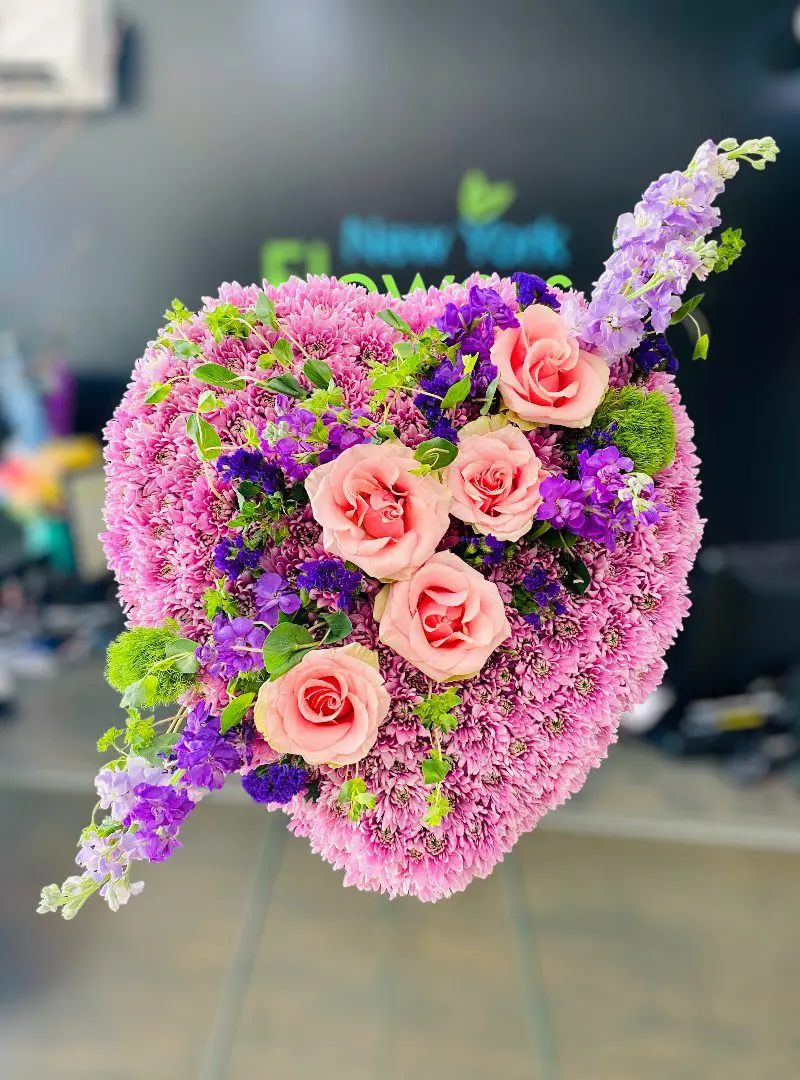 Heart-shaped floral arrangement with pink roses and purple accents on a stand.
