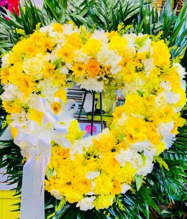 A circular floral wreath composed of YELLOW-WHITE STANDING HEART flowers, accented with a white bow, displayed in front of green foliage.