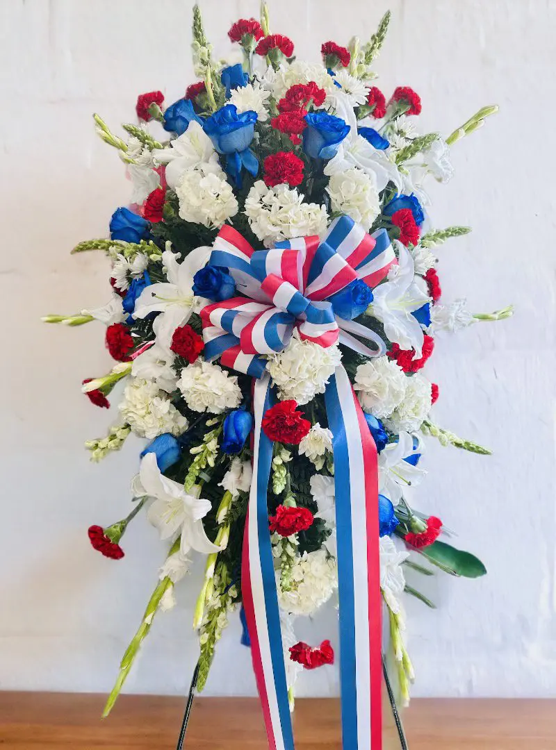 A vibrant floral arrangement featuring a PATRIOTIC STANDING SPRAY with red, white, and blue flowers and a patriotic ribbon displayed against a white background.