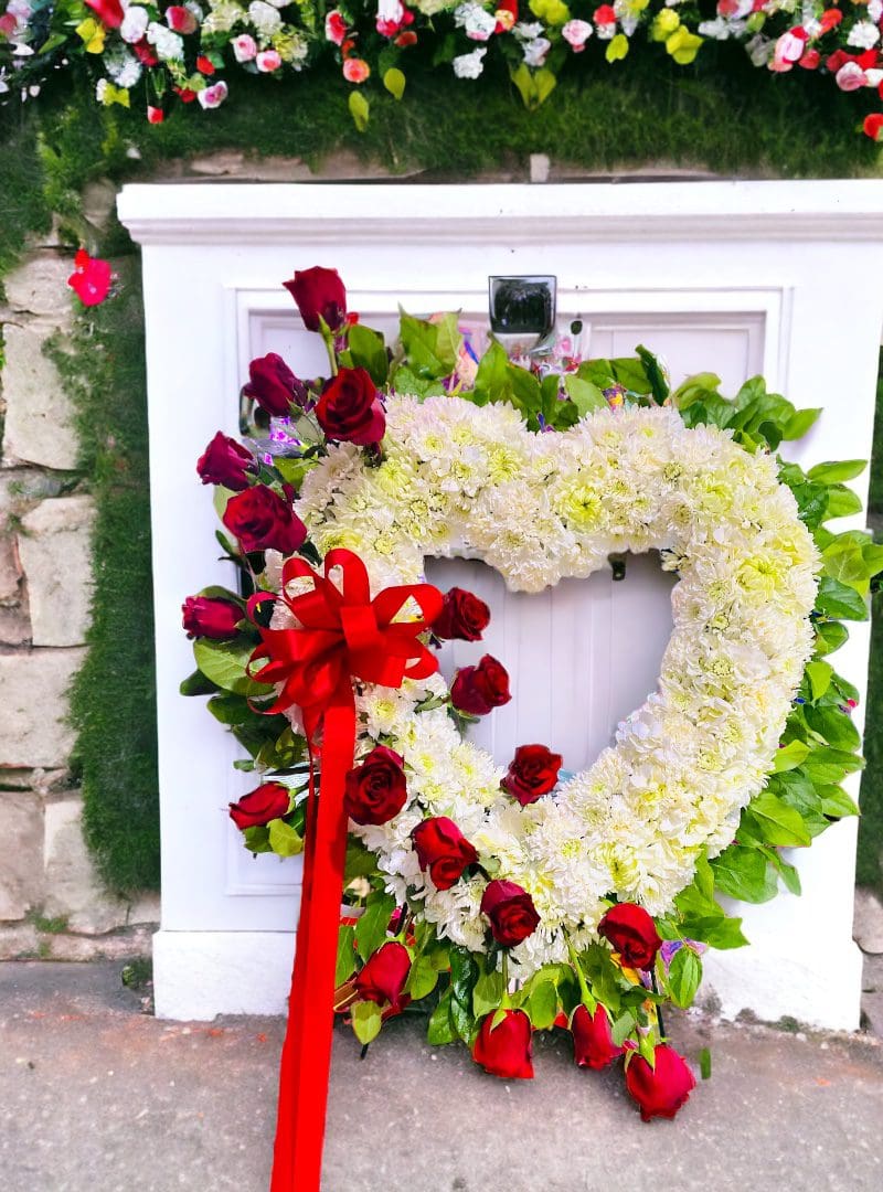A heart-shaped floral wreath with red roses and a large red bow on a white display stand, with a backdrop of assorted flowers.
