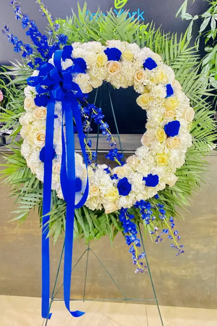 A floral wreath with white and yellow roses, blue flowers, and a blue ribbon displayed on a stand.