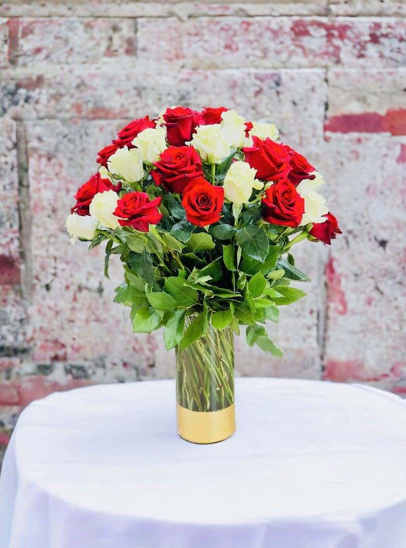 A Red and White Roses Vase Arrangement on a table with a white cloth, set against a brick wall.