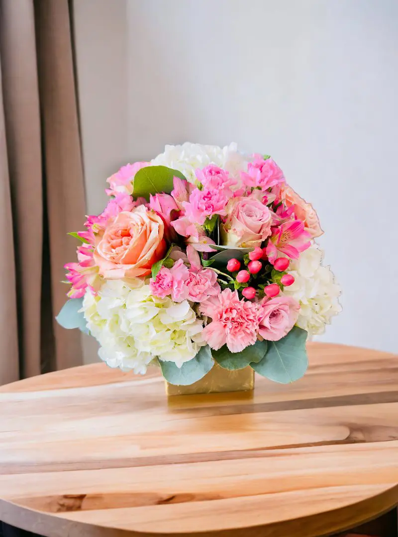 A floral arrangement featuring pink roses and white hydrangeas in a gold vase on a wooden table.