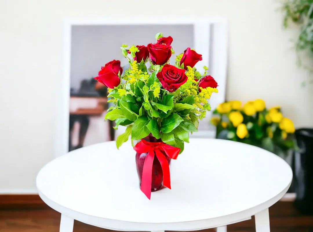 A 12 Long Stem Red Rose in Red vase and green leaves tied with a red ribbon on a white table.