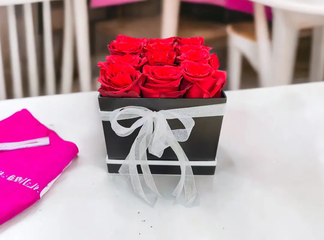 A 12 Red Roses in Black Square Box filled with red roses and tied with a silver ribbon on a white table.