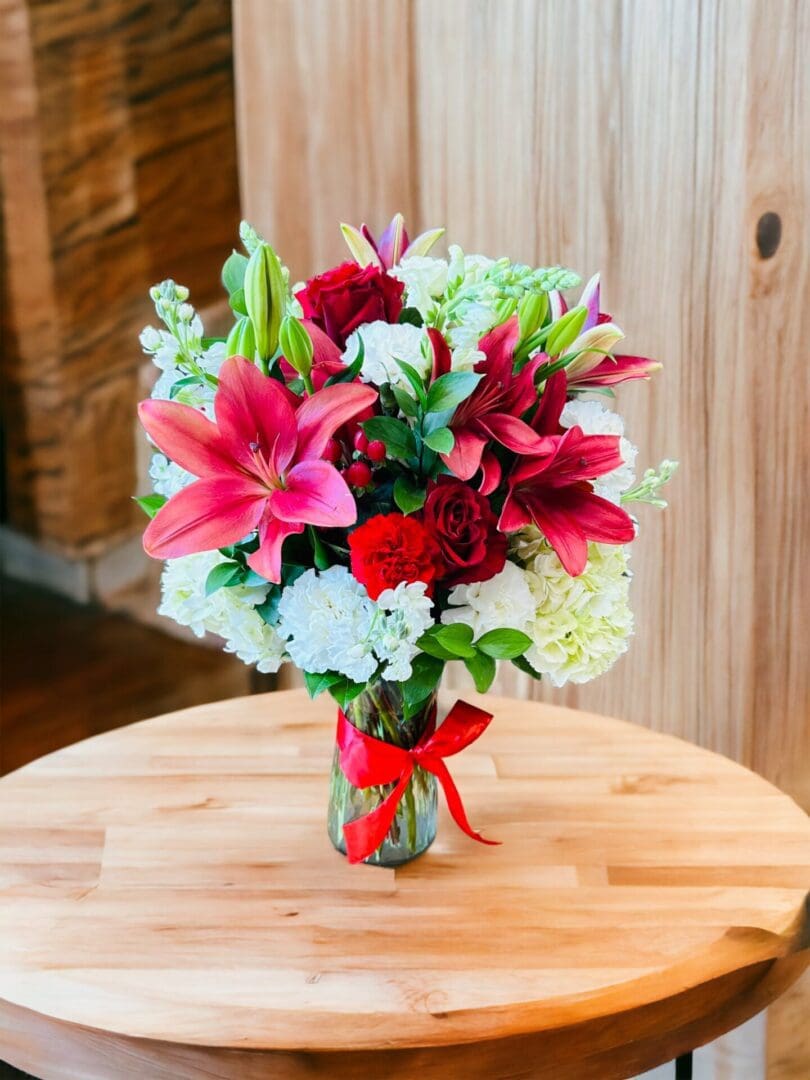 A vibrant Sweetheart Wrapped Bouquet of flowers with lilies, roses, and other blooms arranged in a glass vase on a wooden table.
