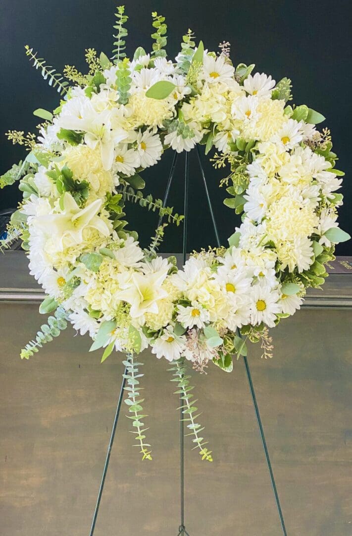 A white floral wreath with green accents displayed on a black background using a metal stand.