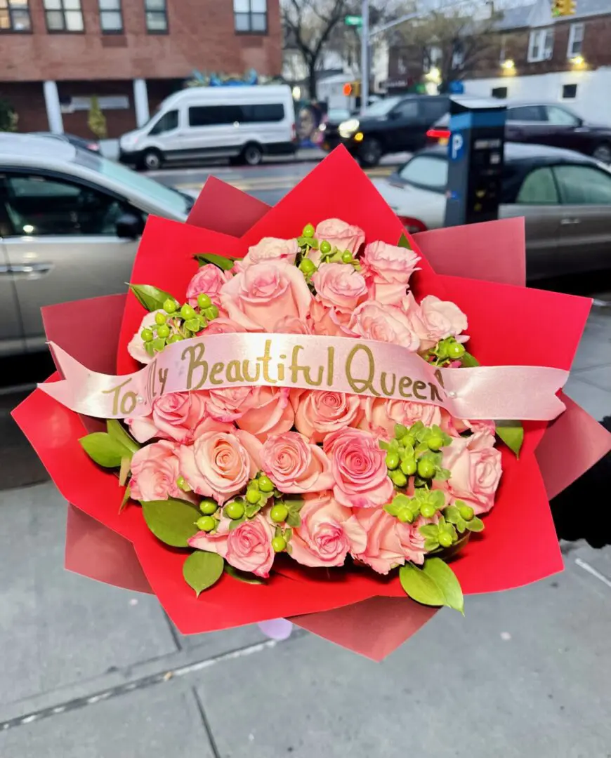 A bouquet of 25 Pink Roses Luxury Bouquet and greenery with a ribbon reading "to my beautiful queen," held up against an urban street background.