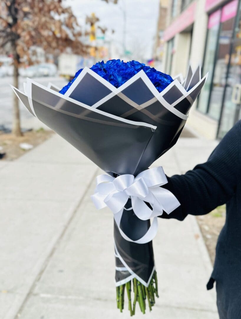 A person holding a bouquet of blue roses wrapped in black and white paper with a white ribbon.