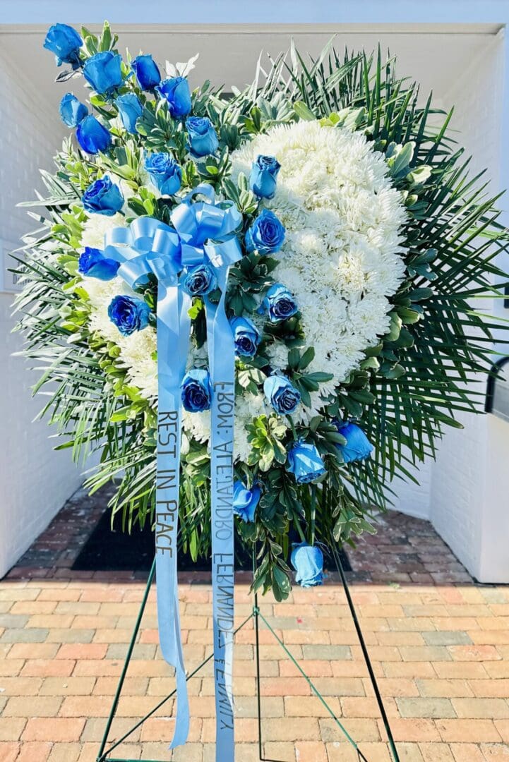 A floral tribute featuring white and dyed blue flowers arranged in a heart shape with a "rest in peace" ribbon.